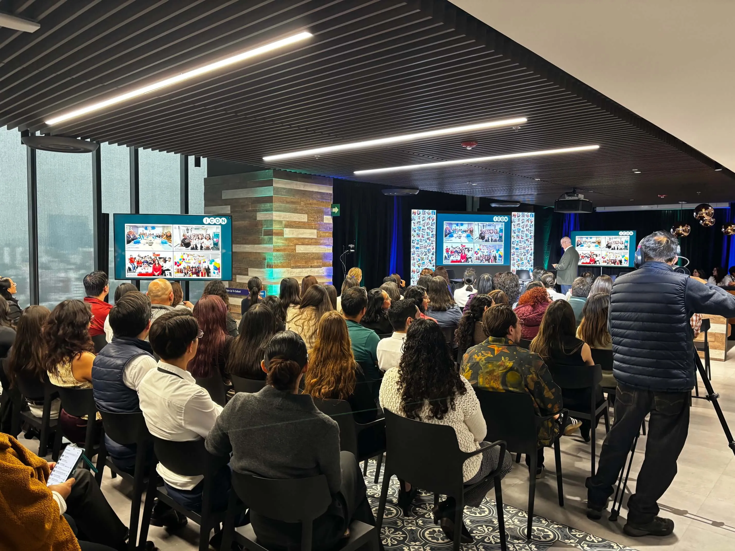 ICON colleagues in the Mexico City cafeteria watching the global program on room displays while a camera operator covers the presenter.